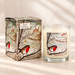 A scented candle in a clear glass jar with cream wax and a winter woodland label, shown next to its matching illustrated box. The label and box feature a robin bird, bare trees, deer, and the text 'Plum Pudding'.